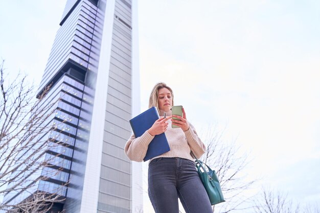 Portrait of a young female student with notebooks and books in hand looking at her smart phone