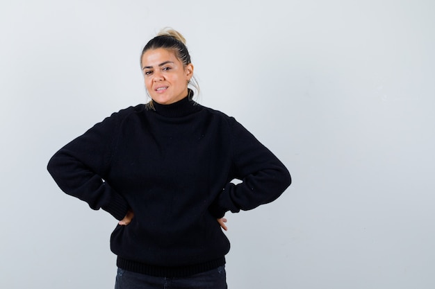 Portrait of young female with hands on hip in black turtleneck sweater and looking cheery front view