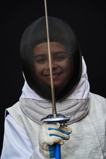 Portrait of young fencer boy wearing white fencing costume and mask and holding the sword in front of her. Isolated on black background