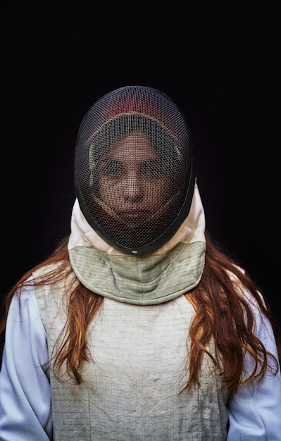 Portrait of a young fencer wearing white fencing costume and\
mask and holding the sword in front of her. isolated on black\
background