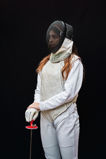 Portrait of a young fencer wearing white fencing costume and\
mask and holding the sword in front of her. isolated on black\
background