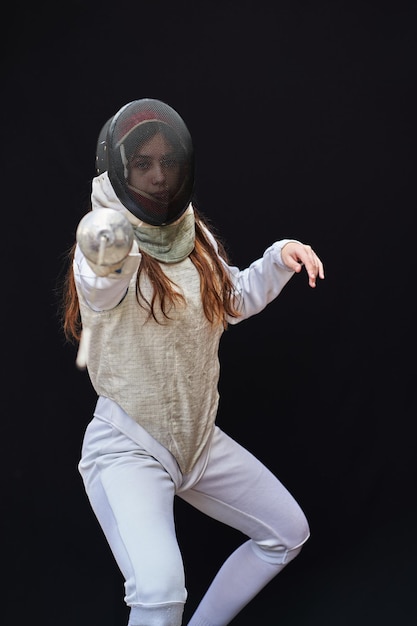 Portrait of a young fencer wearing white fencing costume and\
mask and holding the sword in front of her. isolated on black\
background