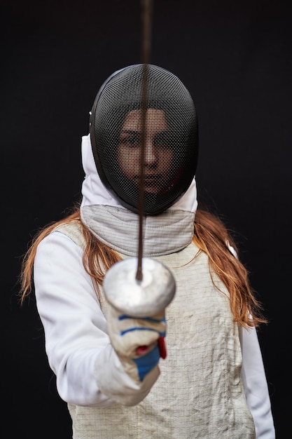 Portrait of a young fencer wearing white fencing costume and\
mask and holding the sword in front of her. isolated on black\
background