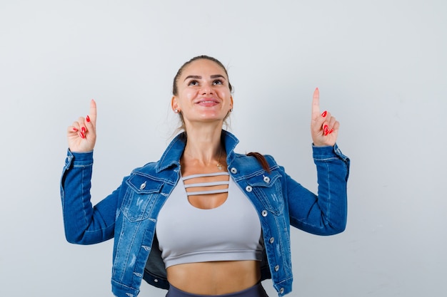 Portrait of young fit female pointing up in top, denim jacket and looking blissful front view