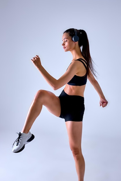 Portrait of young fitness sporty woman in black sportswear and headphones posing, training, working out, doing stretching exercising for legs looking aside isolated on gray color background