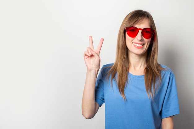 Portrait of a young friendly woman with a smile in a casual blue t-shirt, red glasses, makes a greeting gesture, two fingers up on light. Emotional face