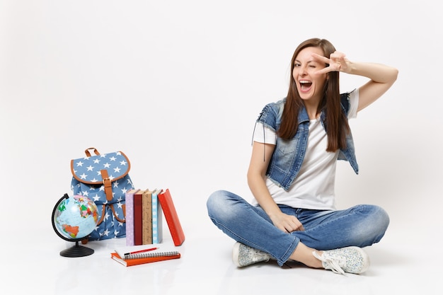 Portrait of young funny joyful woman student in denim clothes showing victory sign sitting near globe, backpack, school books