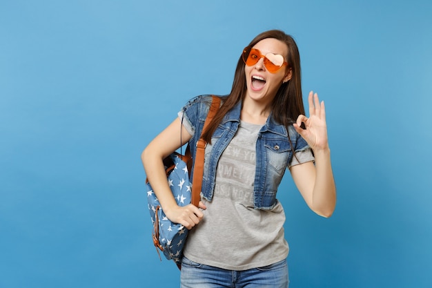 Portrait of young funny joyful woman student with backpack in denim clothes, orange heart glasses showing OK sign isolated on blue background. Education in university. Copy space for advertisement.