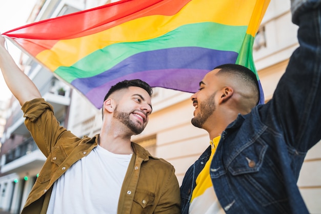 Portrait of young gay couple embracing and showing their love with rainbow flag at the street. LGBT and love concept.