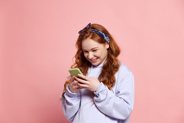 Portrait of young girl in headphones reading text message and
smiling isolated over pink studio background