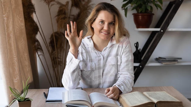 Portrait of a young girl professor sitting at a wooden table\
with books and textbooks at home by the window a woman is preparing\
for a video conference and waves her hand at the camera