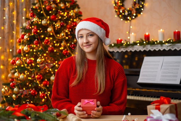 Portrait of a young girl in Santa hat wrapping presents for Christmas for friends and relatives looking to the camera Young smiling girl in decorated living room