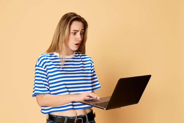 Portrait of young girl in striped shirt styanding with laptop against yellow studio background