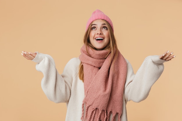 Portrait of young girl wearing winter hat and scarf smiling and looking upward with arms wide open isolated on beige