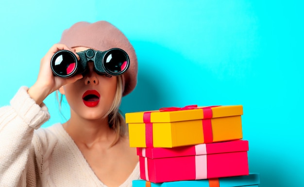 Portrait of a young girl in white sweater with binocular and gift boxes on blue background