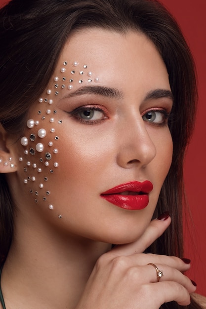 Ritratto di una giovane ragazza con trucco creativo, scintillii e strass sul viso, ritratto di bellezza su uno sfondo rosso.