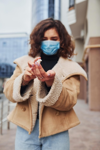 Ritratto di giovane ragazza con capelli ricci in maschera protettiva che usa antisettico all'aperto vicino all'edificio aziendale al momento della quarantena concezione del coronavirus