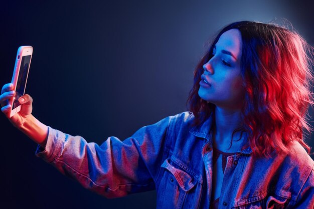 Portrait of young girl with curly hair that makes selfie by using phone in red and blue neon in studio