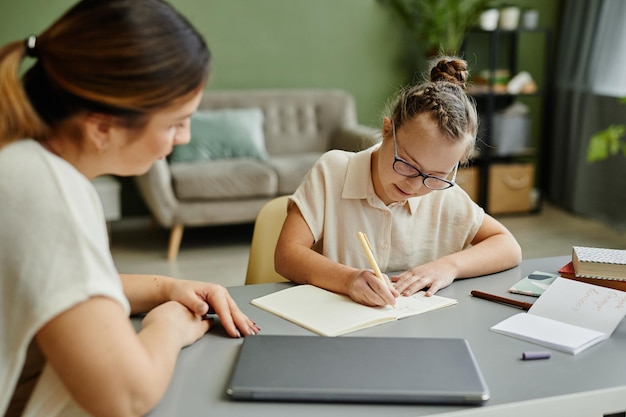 Portrait of young girl with down syndrome studying at home with female tutor or mother helping