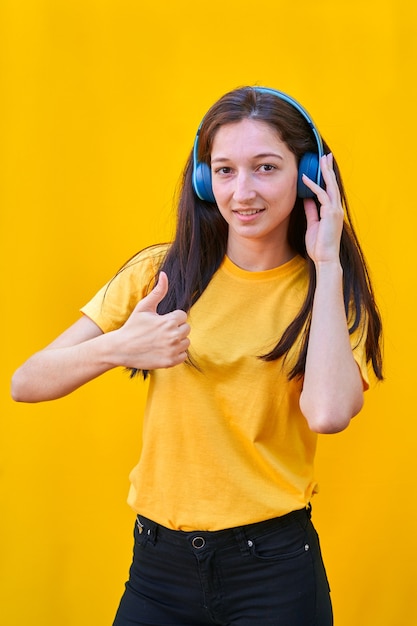 긴 갈색 머리 노란색 tshirt와 검은 색 청바지가 오른손의 엄지 손가락으로 그녀의 파란색 헤드폰으로 음악을 듣고 어린 소녀의 초상화