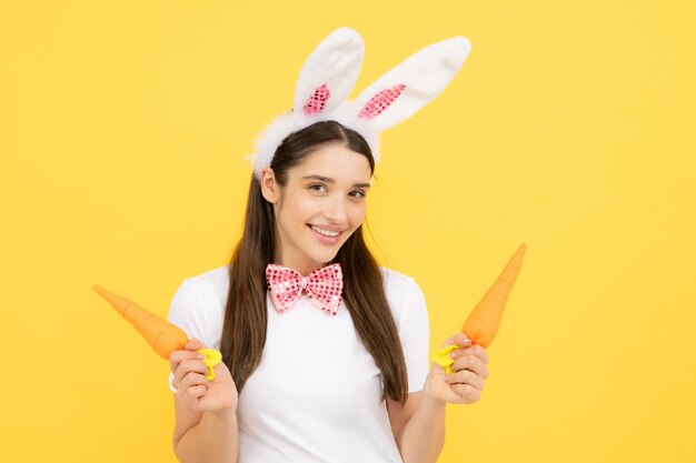 Portrait of young girl with rabbit bunny ears isolated on yellow background Easter bunny woman with toy carrot looks fun