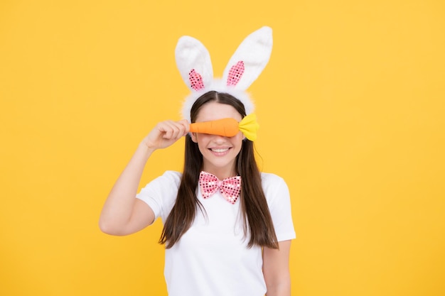 Portrait of young girl with rabbit bunny ears isolated on yellow background Easter bunny woman with toy carrot looks fun