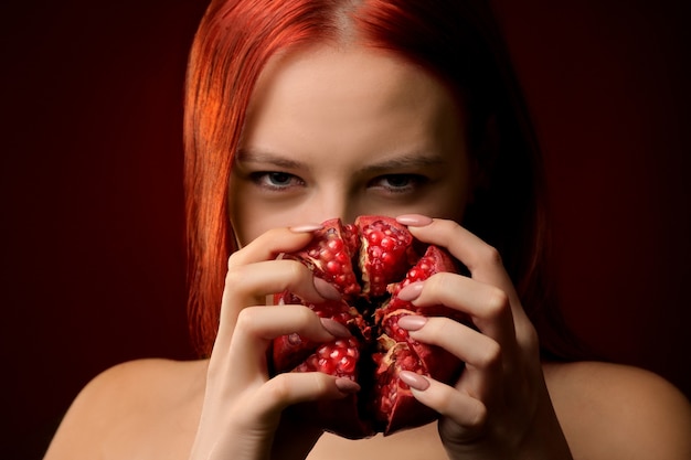 Portrait of a young girl with red hair and pomegranate fruit covering her face on a red background