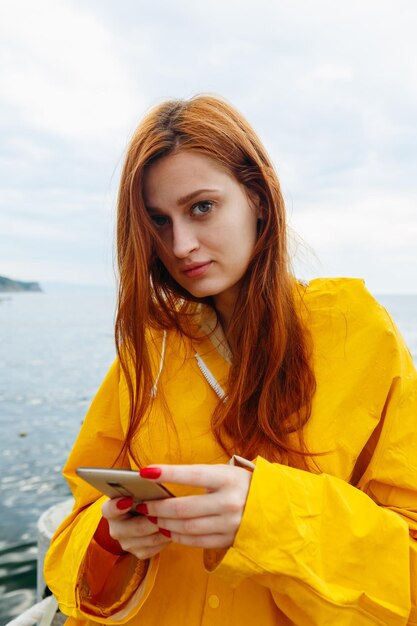 Portrait of young girl with red hair wearing bright coat and posing on ocean looking at camera