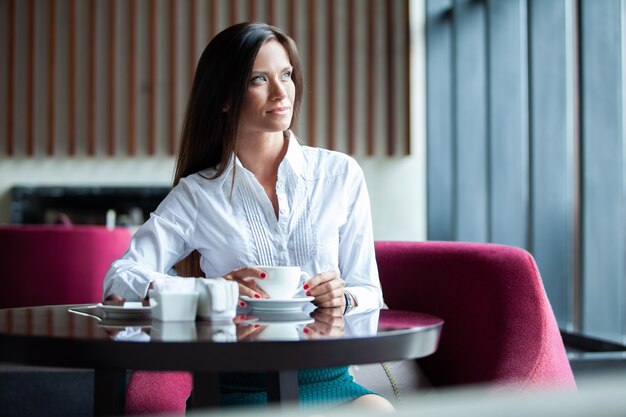 Portrait of young gorgeous female drinking tea and looking with smile out of the coffee shop window while enjoying her leisure time, nice business woman lunch in modern cafe during her work break.