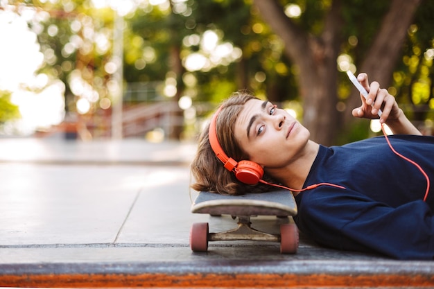 Portrait of young guy in orange headphones lying on skateboard with cellphone in hand while dreamily looking in camera at skatepark