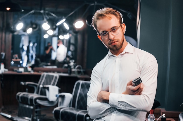 Portrait of young guy in white clothes that standing indoors in barber shop and holding clipper