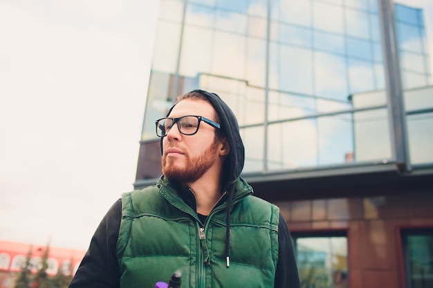 Portrait of young guy with large beard in glasses vaping an electronic cigarette opposite urban background.