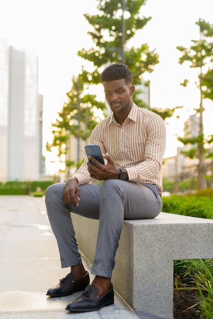 Portrait of young handsome African businessman sitting outdoors in city using mobile phone