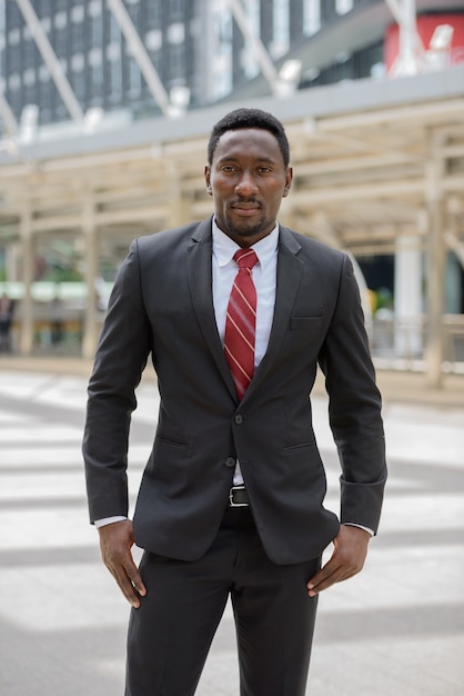 Portrait of young handsome African businessman in suit against view of modern building in the city outdoors