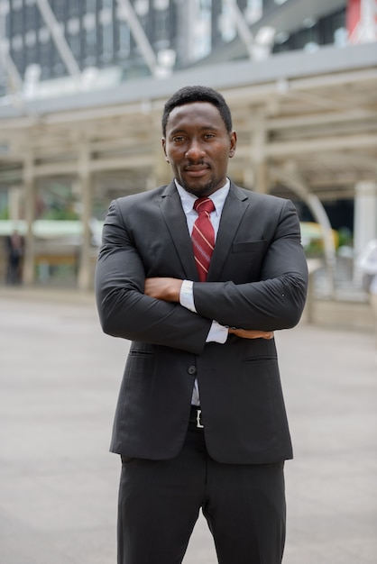 Portrait of young handsome African businessman in suit against view of modern building in the city outdoors