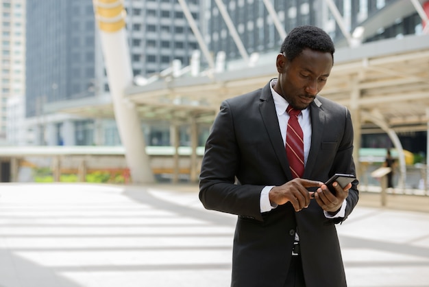 Portrait of young handsome African businessman in suit against view of modern building in the city outdoors