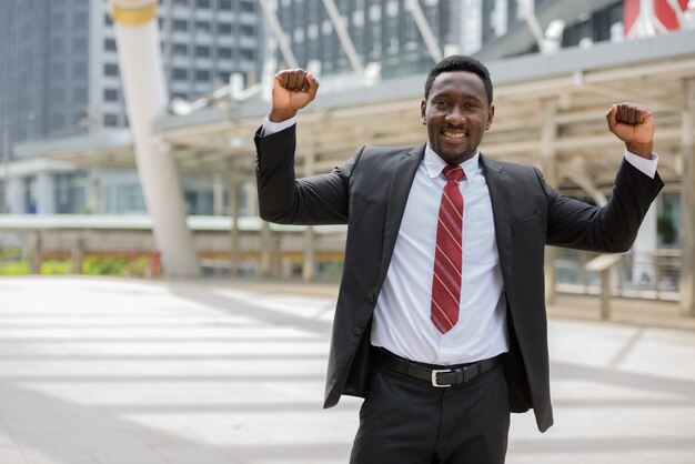 Portrait of young handsome African businessman in suit against view of modern building in the city outdoors