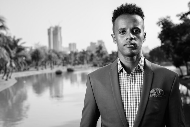 Portrait of young handsome African businessman wearing suit while relaxing at the park in black and white