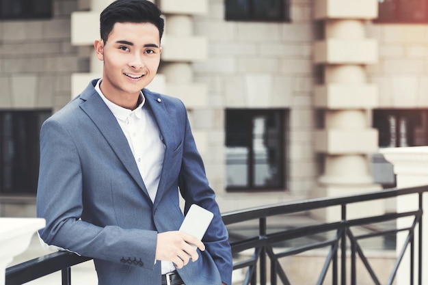 Портрет молодого красивого азиатского бизнесмена в костюме, стоящего на улице и держащего смартфон. Тонированное изображение