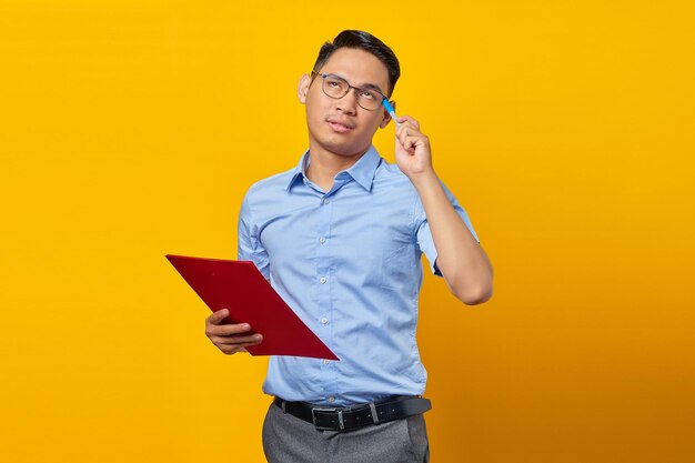 Portrait of young handsome Asian man scratching head with pen thinking of ideas and holding document folder isolated on yellow background