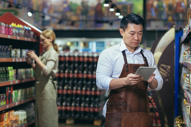 近くのエプロンに立っている若いハンサムなアジア人男性店主スーパー マーケット労働者の肖像画