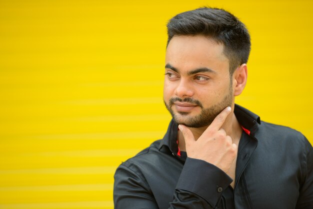 Portrait of young handsome bearded indian businessman against yellow wall