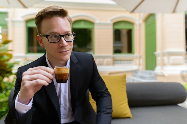 Portrait of young handsome blond businessman in suit relaxing at the coffee shop outdoors