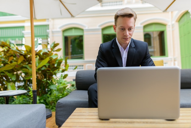 Portrait of young handsome blond businessman in suit relaxing at the coffee shop outdoors