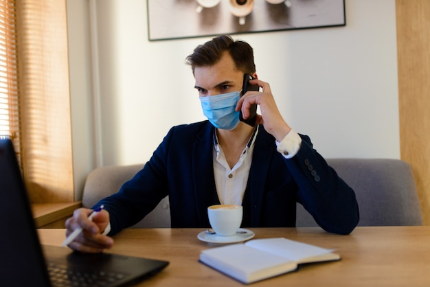 Portrait of young handsome businessman with face mask in quarantine of flu. Photo in caffee with laptop, mobile phone.