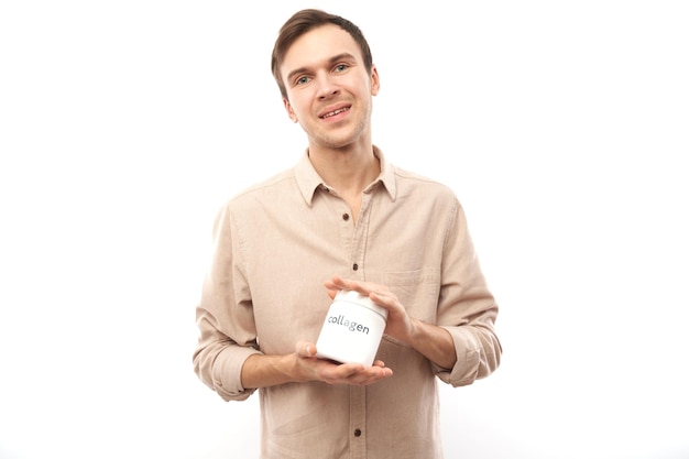 Portrait of young handsome caucasian man showing jar of collagen powder in his hands and smiling isolated on studio white background