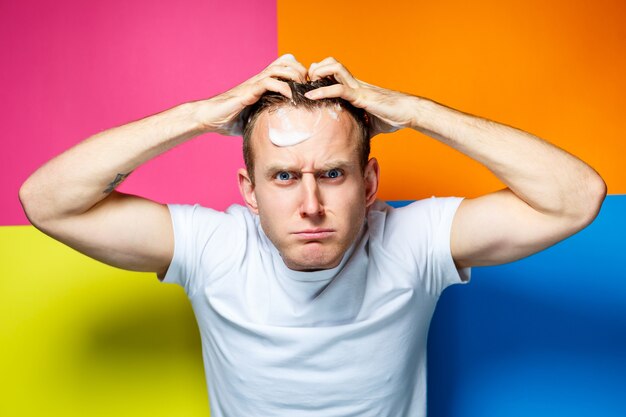 Portrait of a young, handsome, cheerful guy on a multi-colored background, dressed in a white T-shirt, creates a fashionable hairstyle, stupid and positive emotions