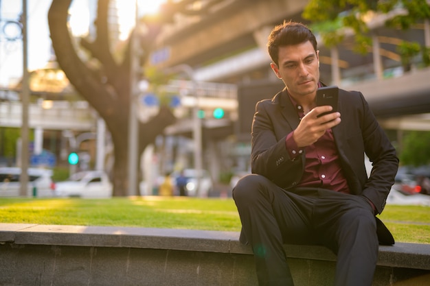 Portrait of young handsome Hispanic businessman sitting and getting fresh air with nature in the city outdoors