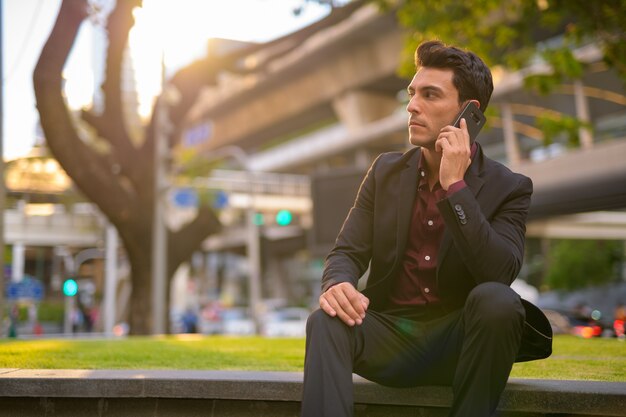 Portrait of young handsome Hispanic businessman sitting and getting fresh air with nature in the city outdoors