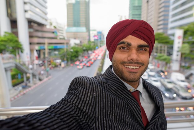 Portrait of young handsome Indian Sikh businessman wearing turban while exploring the city of Bangkok, Thailand